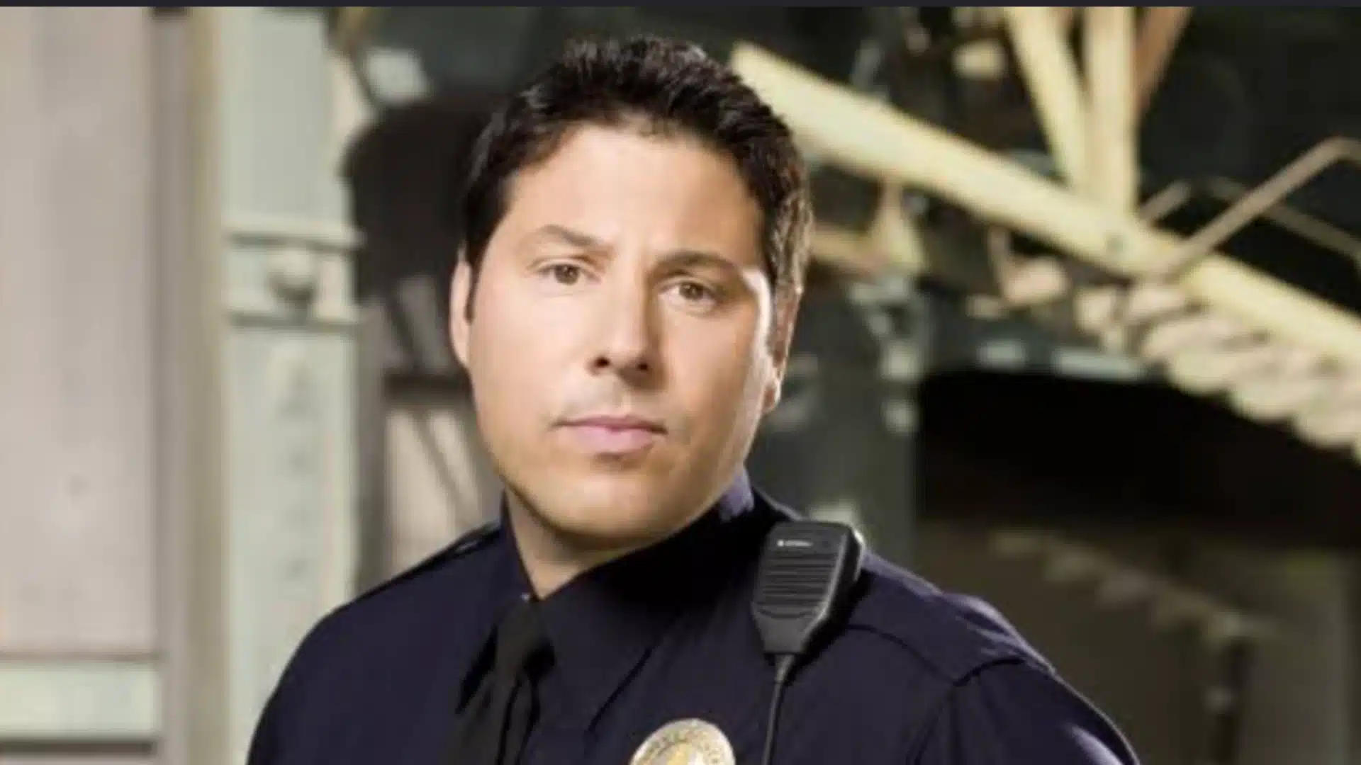 dark-haired man in a police uniform poses indoors with a shoulder radio and blurred industrial beams in the background