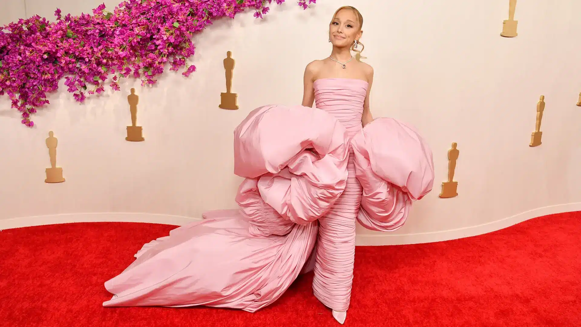 A woman poses on a red carpet in a dramatic strapless pink gown with voluminous ruffles and a long trailing train, against a backdrop of pink flowers and golden statuettes.