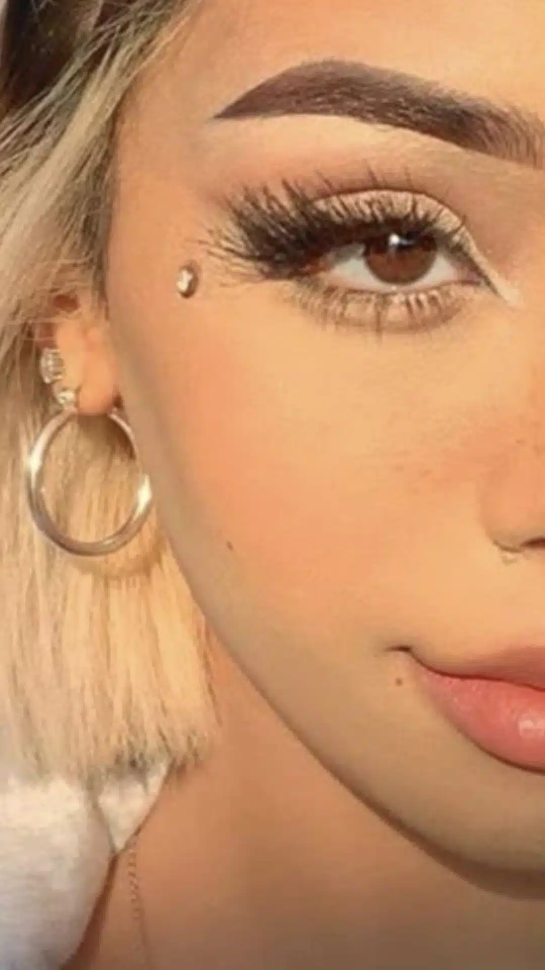 Close-up portrait of a woman with blonde hair, dramatic eyelashes, defined eyebrows, a cheek piercing, and large hoop earrings.