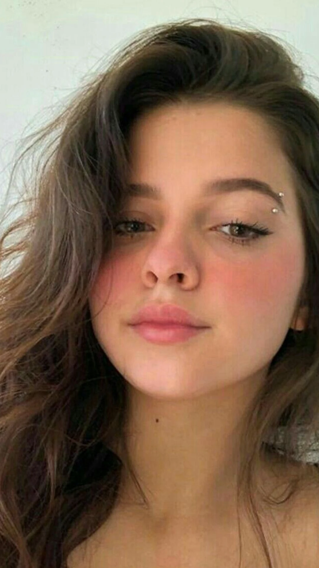 Close-up portrait of a young woman with wavy brown hair, rosy cheeks, winged eyeliner, and an eyebrow piercing.
