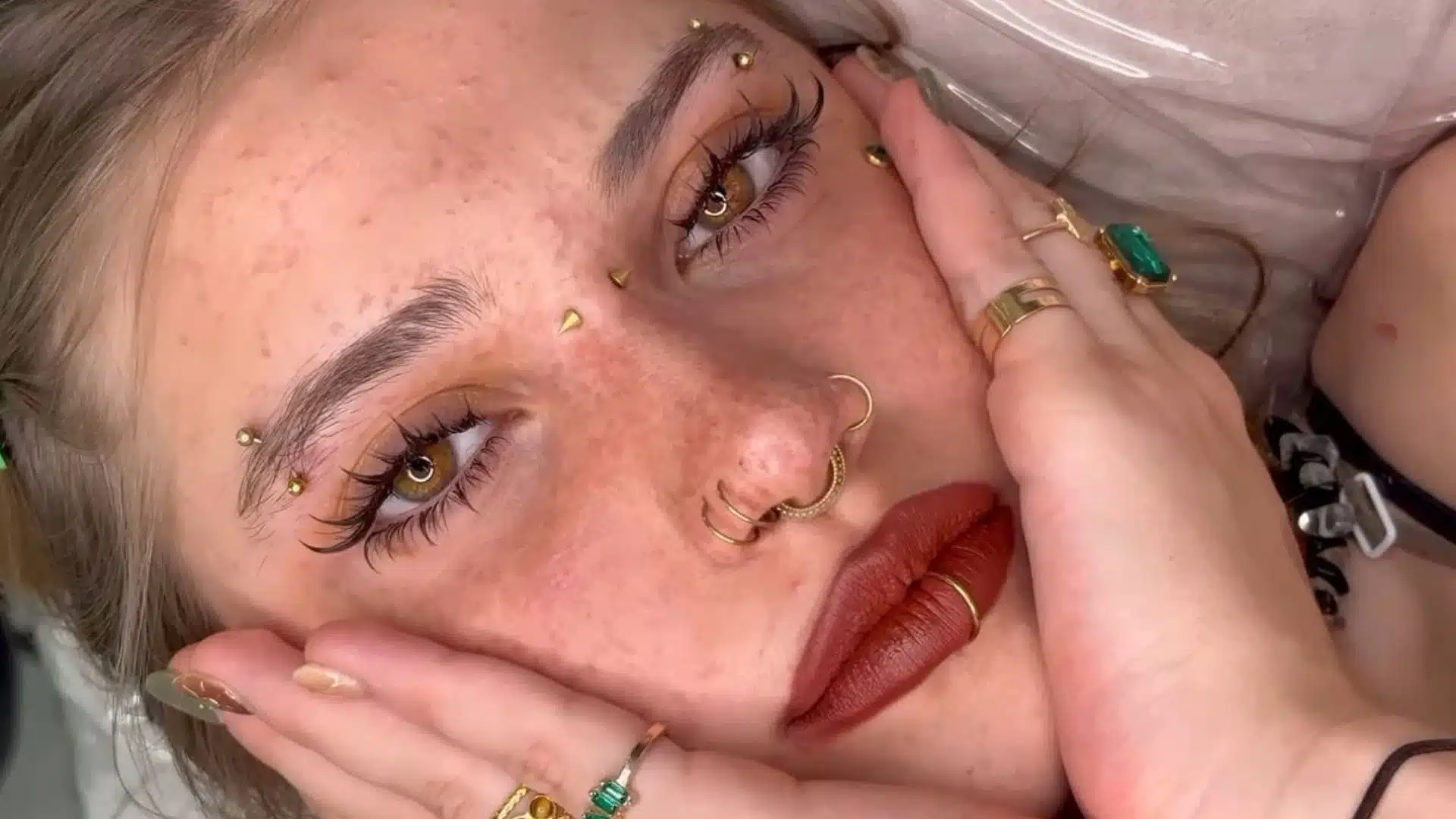 A woman with freckles and piercings reclines on a bed looking relaxed and contemplative
