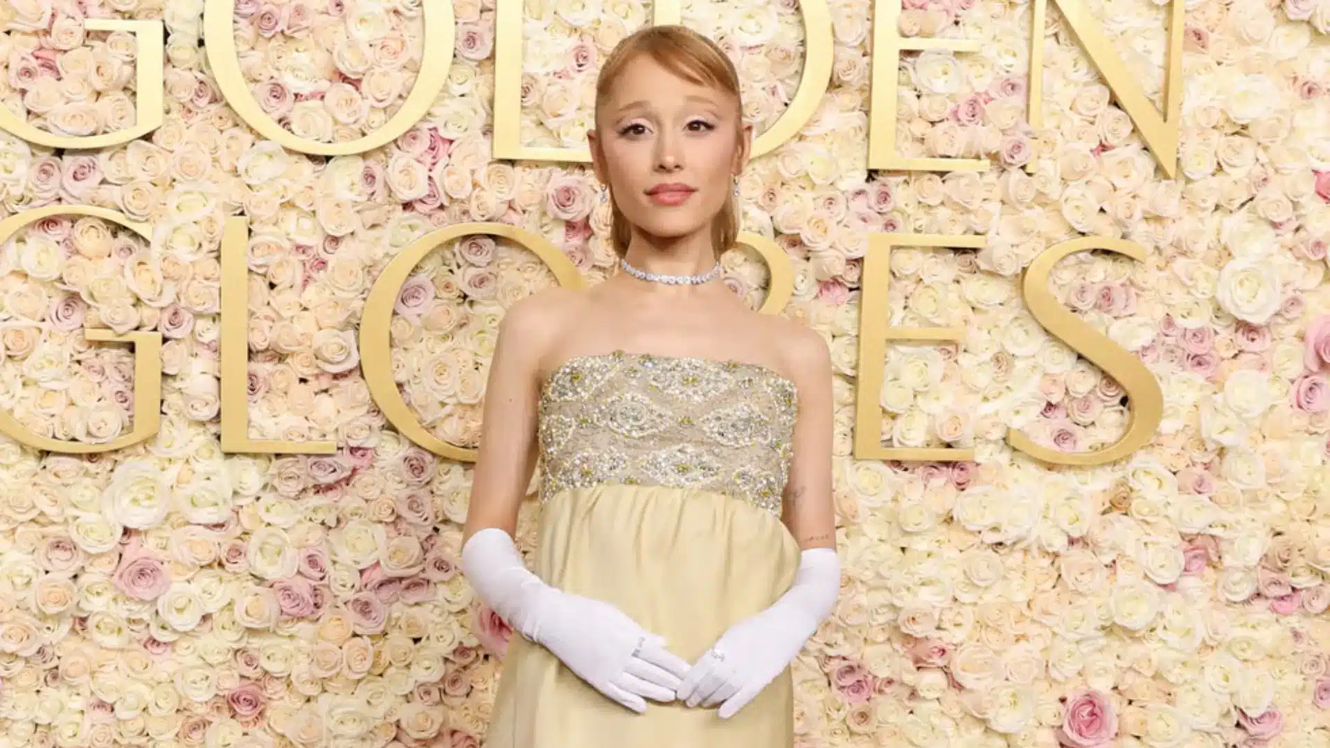 Ariana Grande poses in a strapless embellished gown and white gloves before a wall of pastel roses and gold “Wicked” lettering.