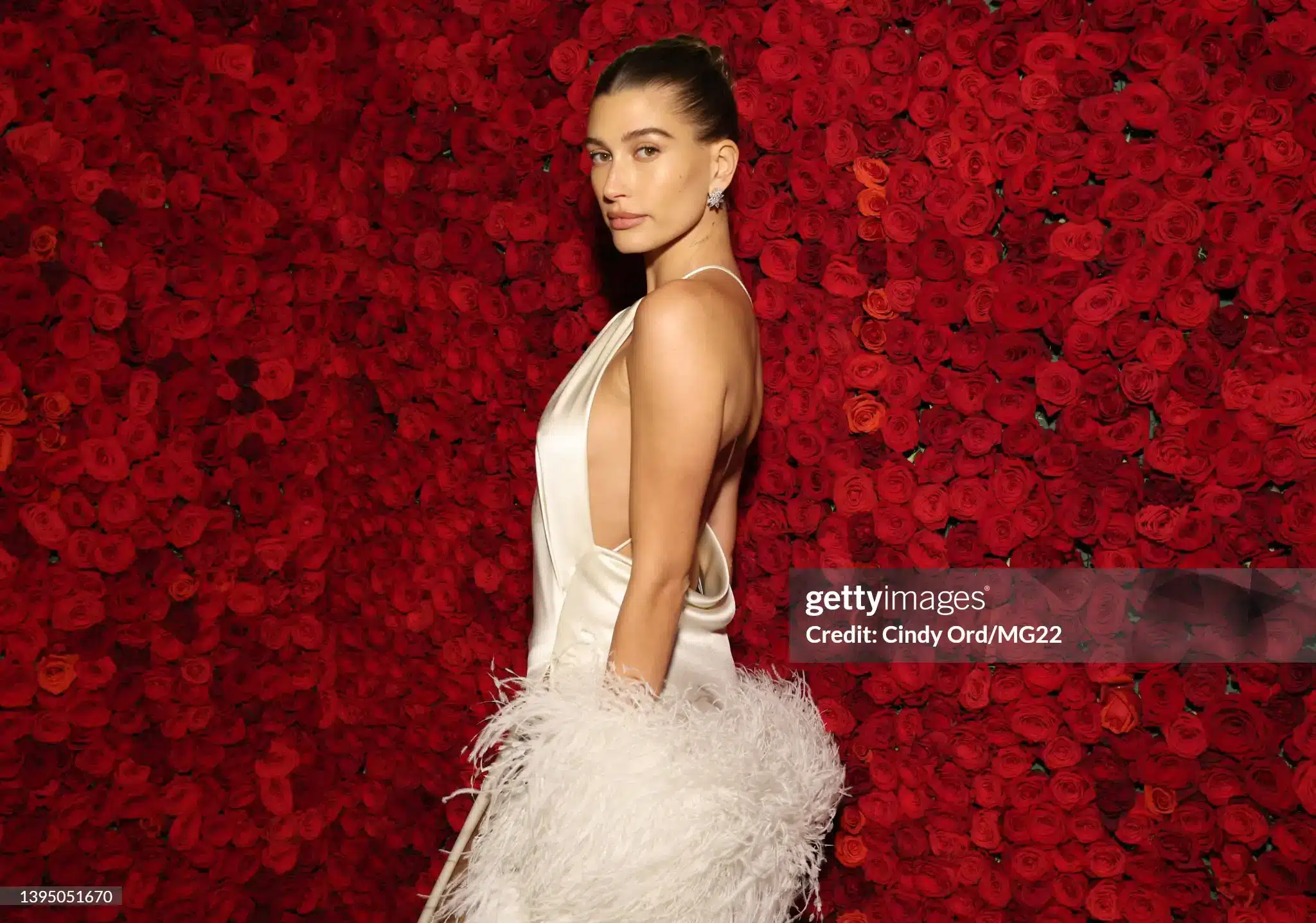Hailey Bieber in a sleek bun posing in a satin gown before a wall of red roses at Met Gala 2022