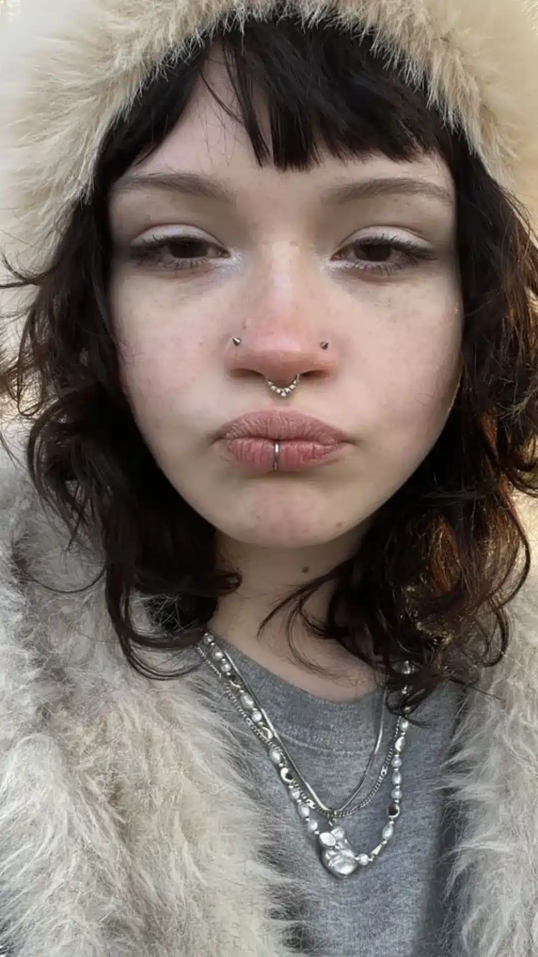 Close-up portrait of a freckled person with dark wavy hair, multiple facial piercings, layered necklaces, and a fluffy gray fur-trimmed hood.