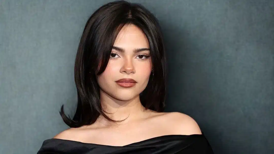 Ariana Greenblatt with dark hair wearing a black dress, posing confidently for the camera.