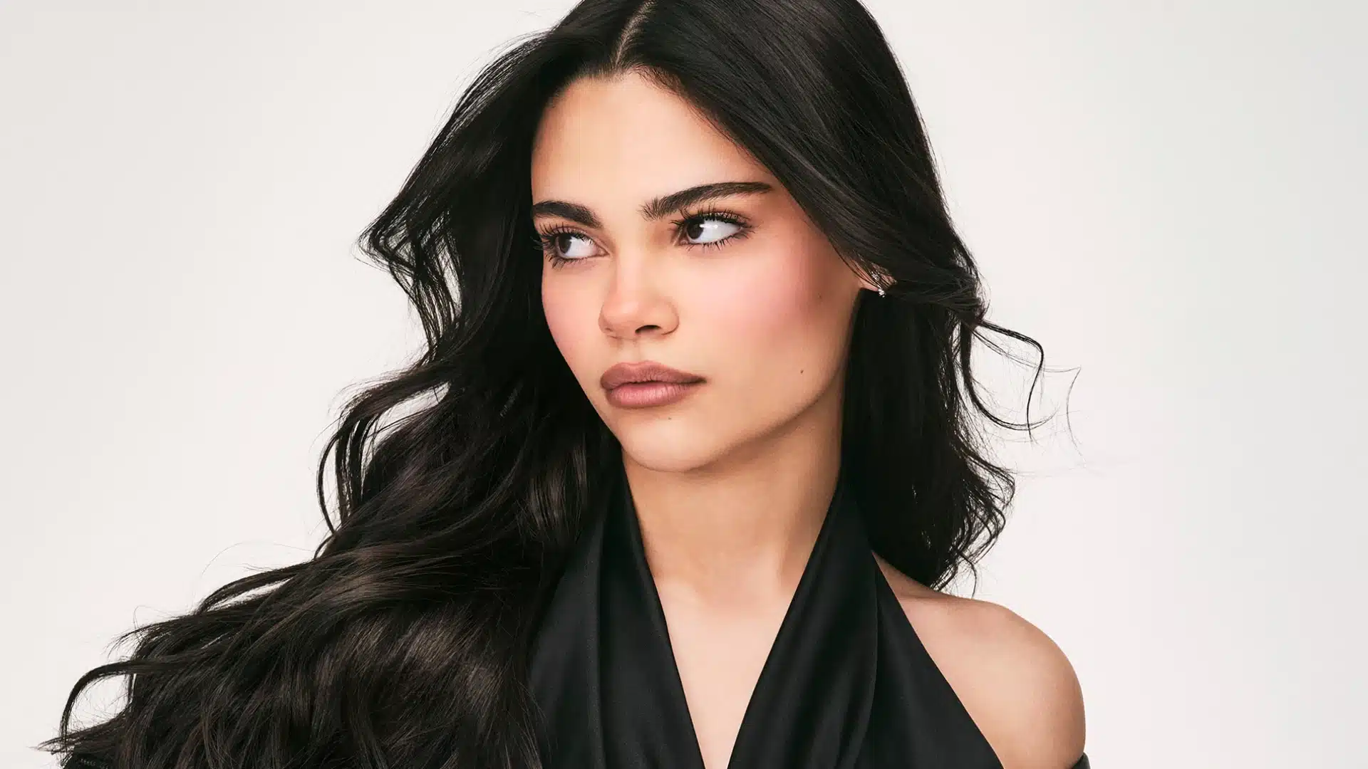 Ariana Greenblatt with long dark hair is wearing an elegant black dress, standing confidently against a neutral background