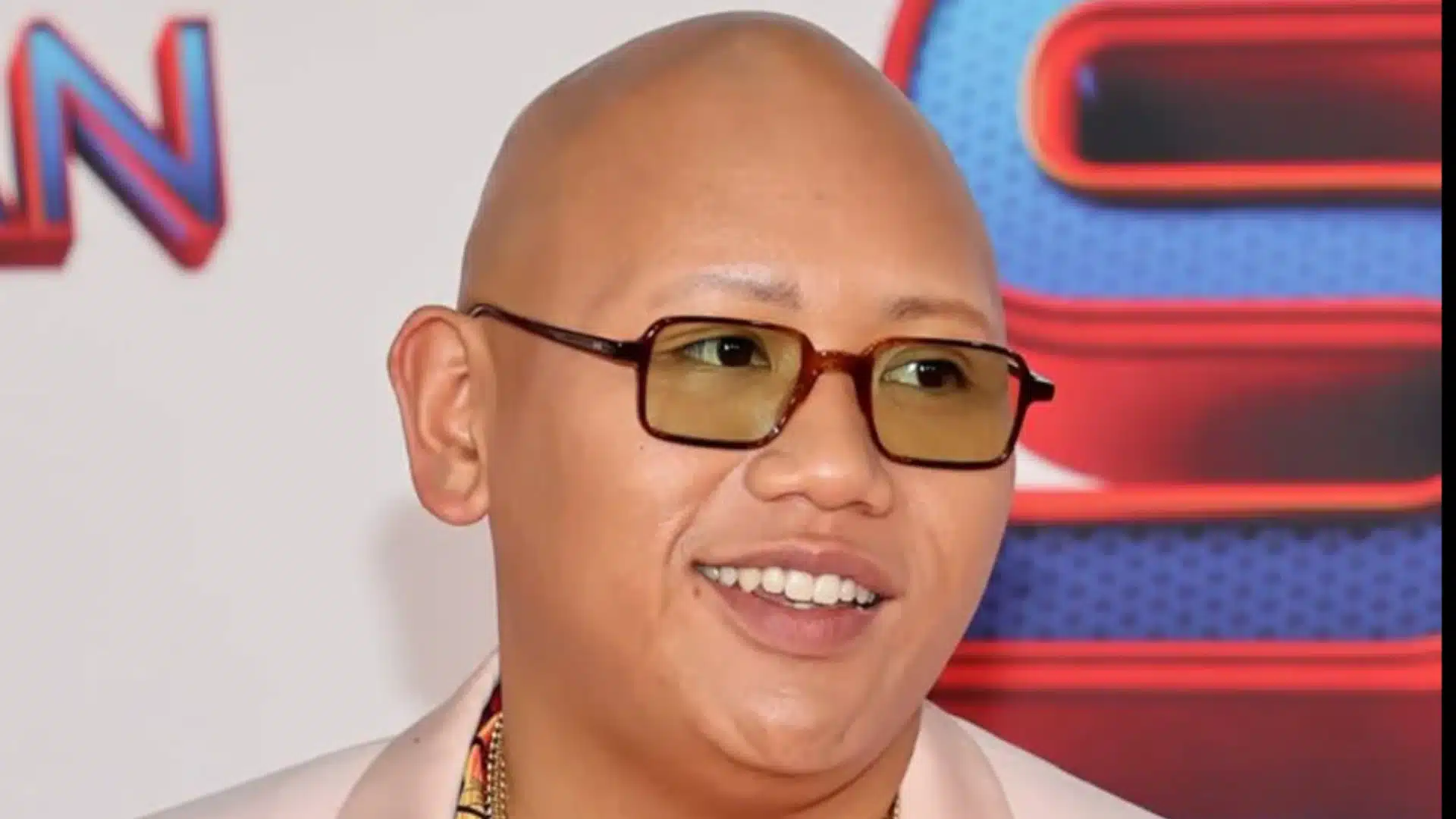 Jacob Batalon smiling with a bald head at a movie premiere, showing his natural appearance due to alopecia.