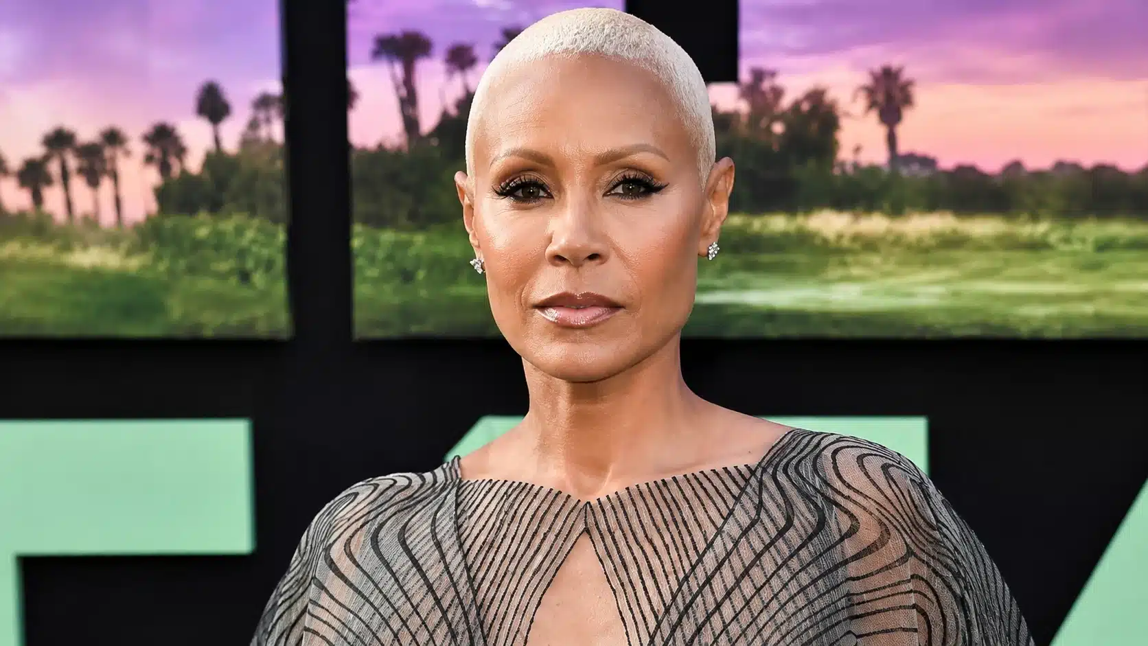 woman with platinum buzz cut and patterned dress poses before a vivid sunset backdrop with palm trees and graphic shapes
