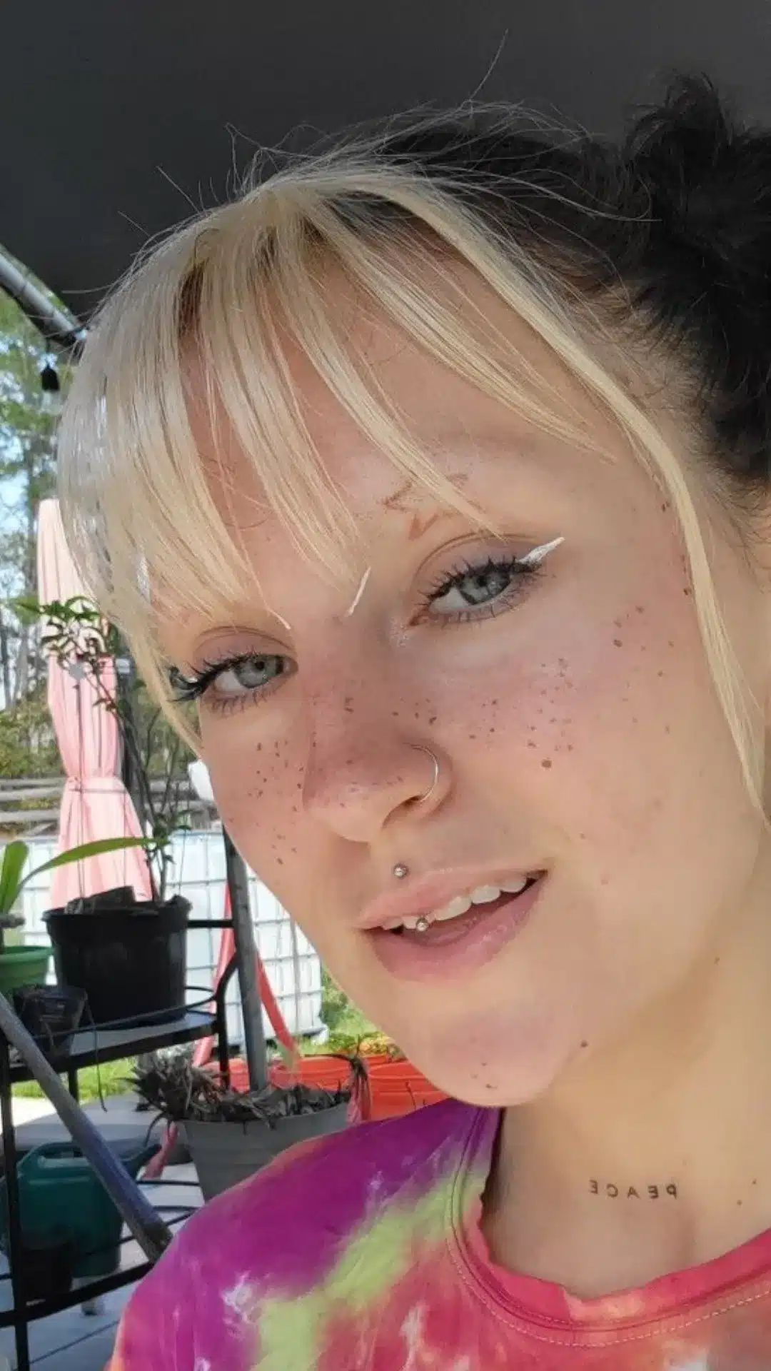 Close-up portrait of a woman with blonde bangs, freckles, winged eyeliner, a nose ring, and a jestrum piercing.