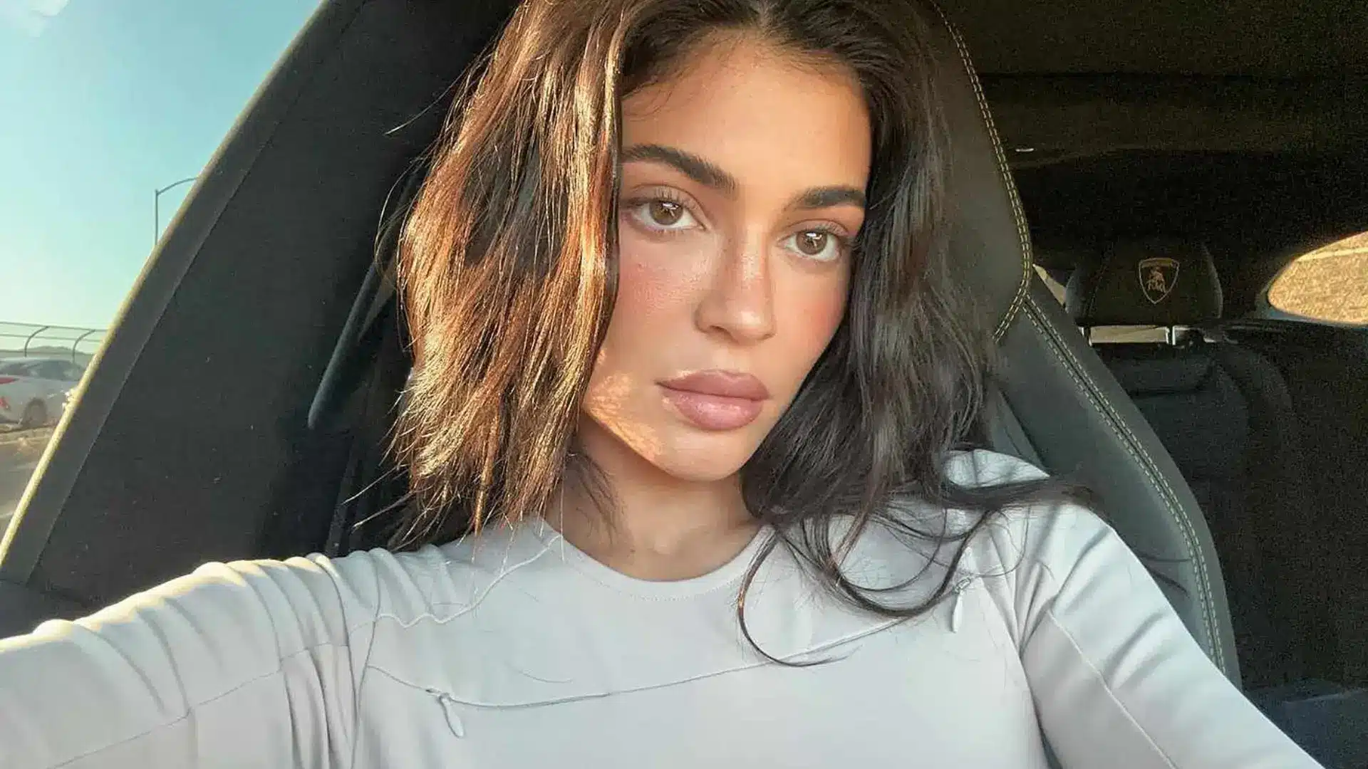 Kylie Jenner with long dark hair and natural makeup taking a selfie inside a car in warm daylight wearing a beige top