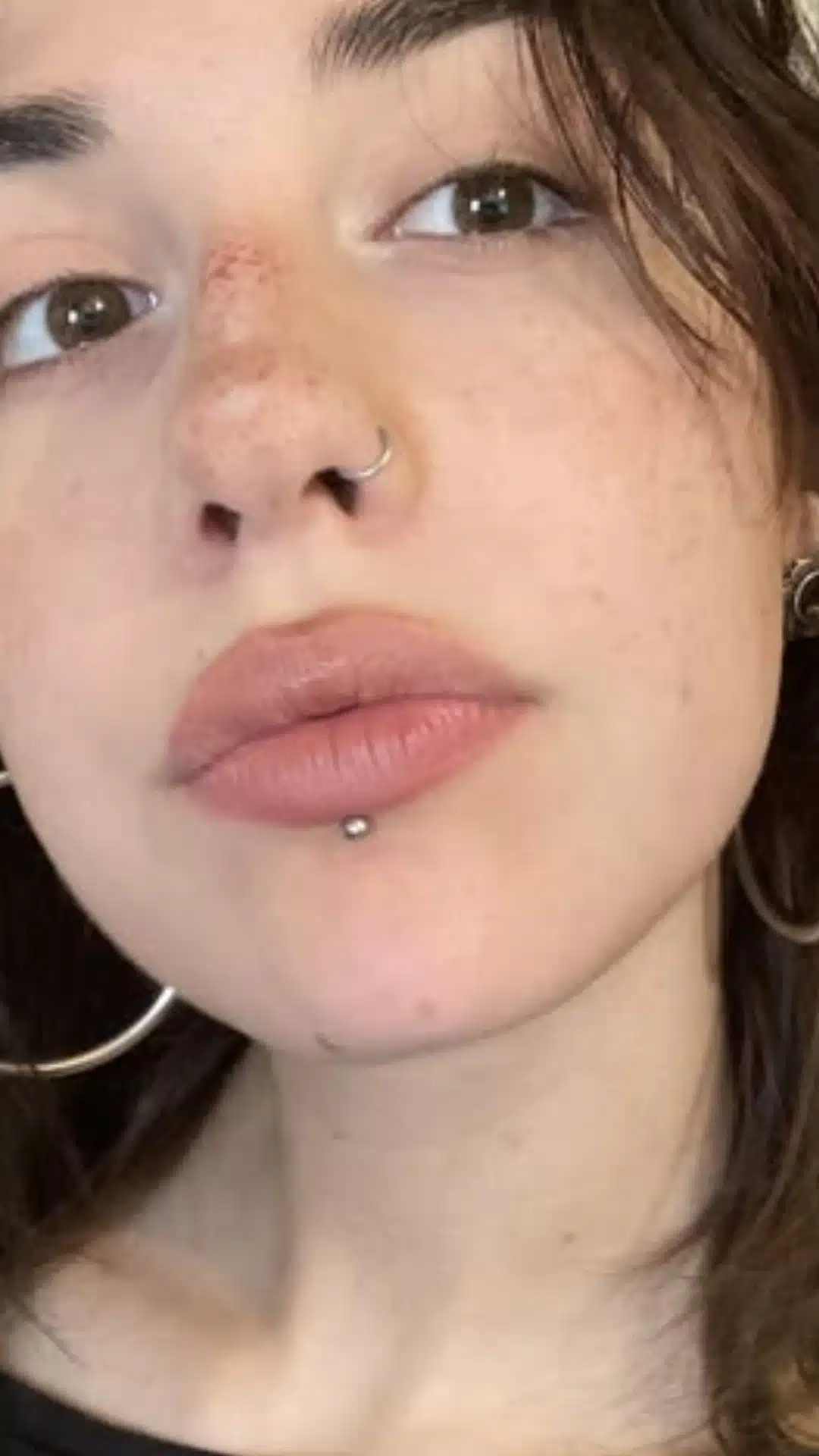 Close-up portrait of a person with brown eyes, freckles, dark hair, a nose ring, large hoop earrings, and a lower-lip stud.