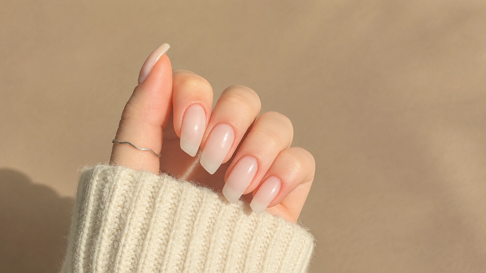 Close-up of glossy nude ombre square nails on a hand in a cream knit sleeve with a thin silver ring under warm sunlight