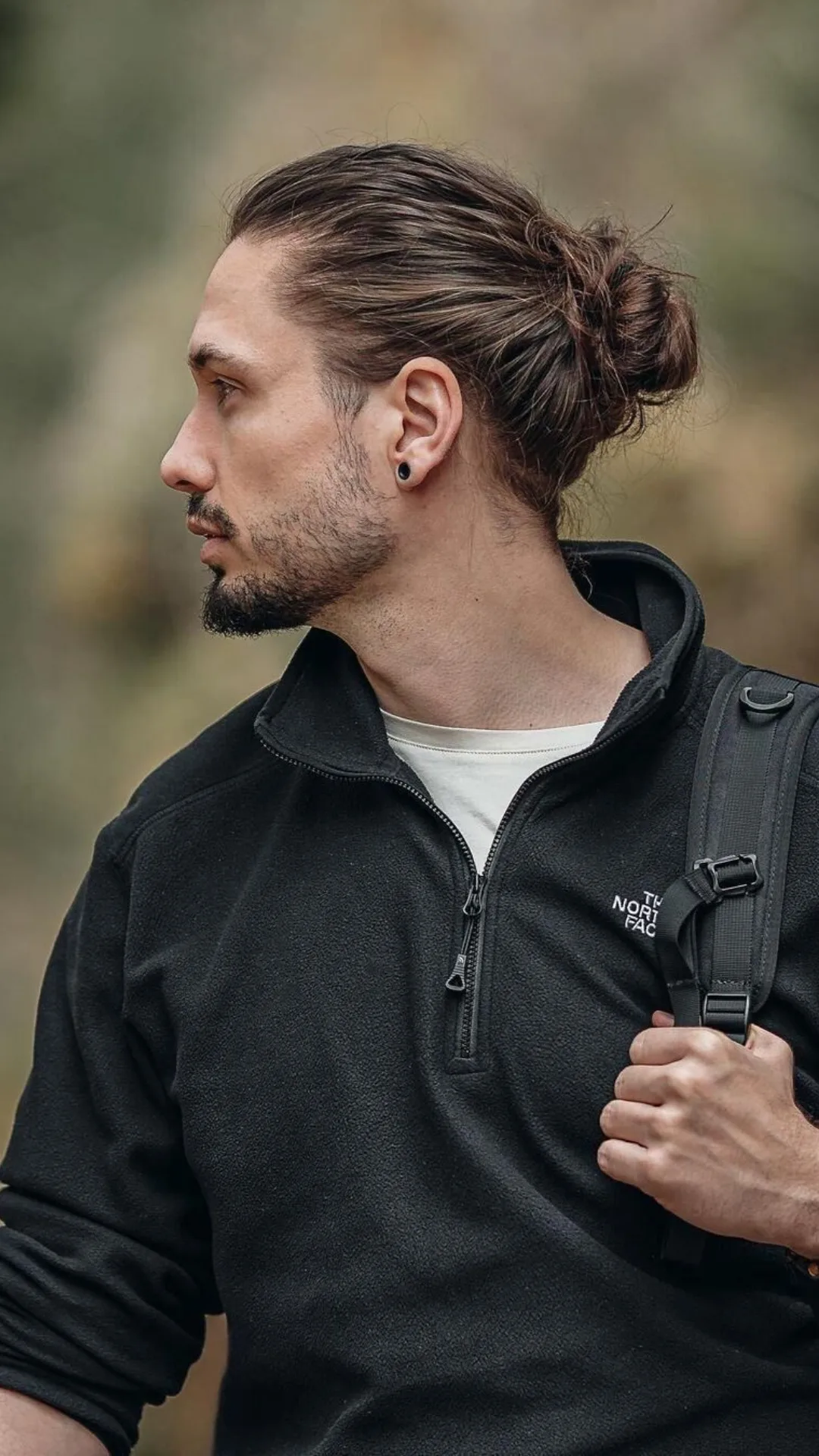 Bearded man with hair tied in a bun wearing a black North Face jacket and backpack, looking to the side outdoors.