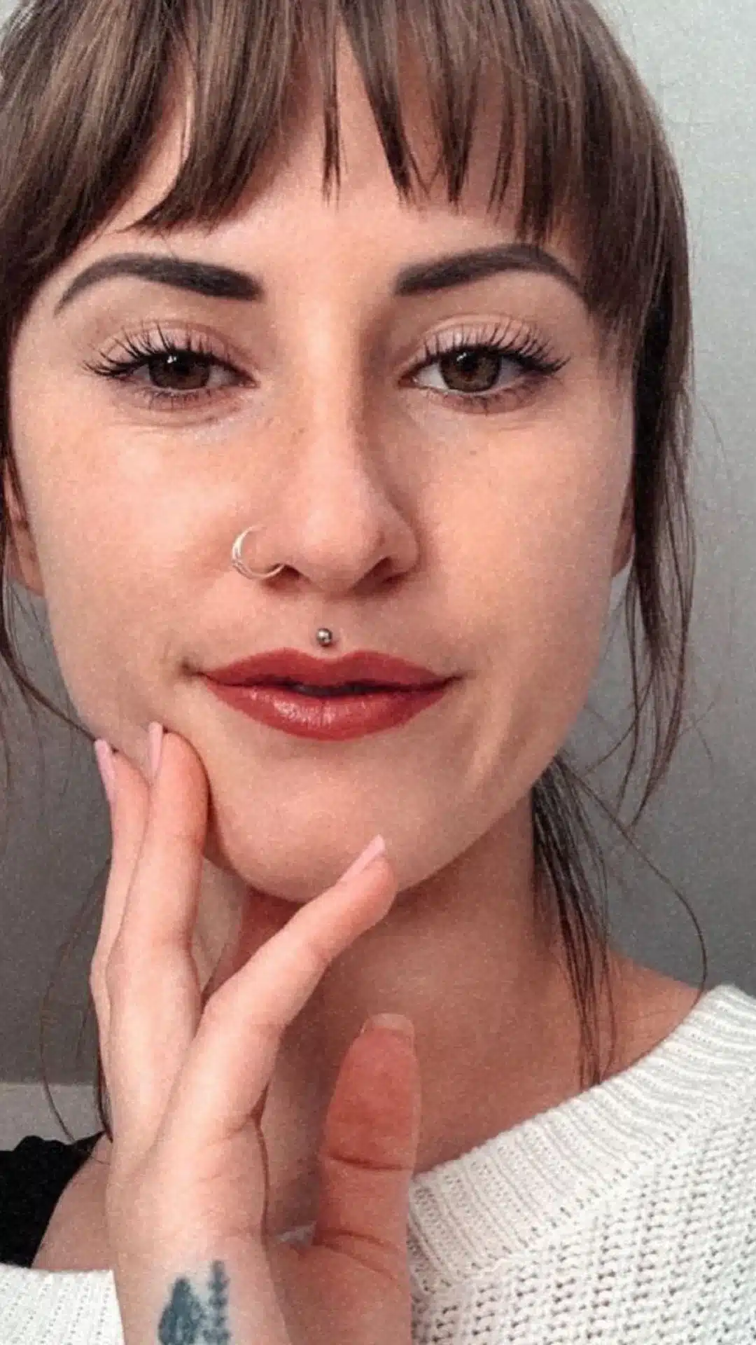 Close-up portrait of a woman with bangs, a silver nose ring, Medusa piercing, red lipstick, and subtle eye makeup.