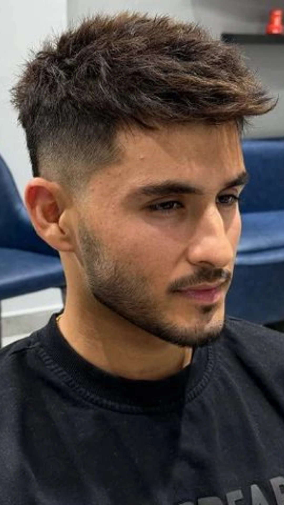 Young man with textured brown hair, a faded haircut, and trimmed beard wearing a black shirt while seated in a salon chair.