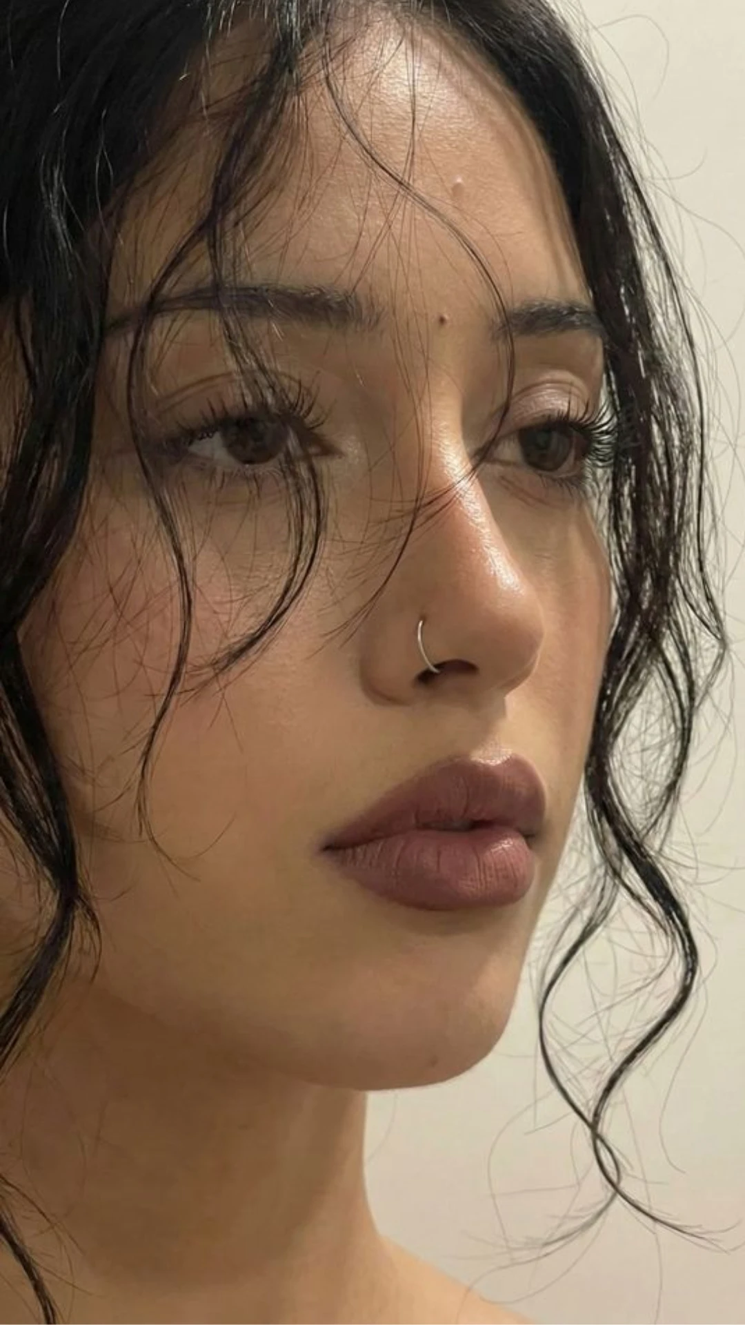 Close-up portrait of a woman with tousled dark hair, long eyelashes, glossy lips, and a small silver nose ring.