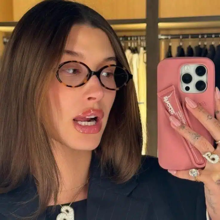 Hailey Bieber wearing oval-frame glasses smiles while taking a selfie, highlighting her fashionable look and modern style.