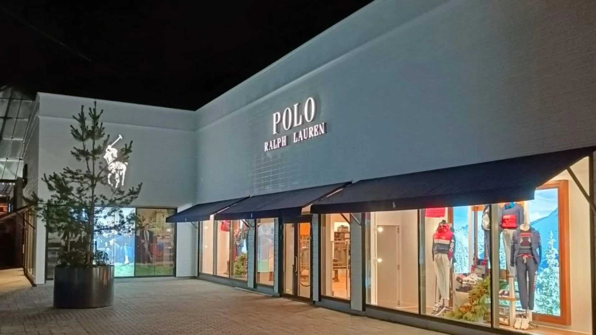 Polo Ralph Lauren storefront with illuminated display windows, mannequins, and a potted evergreen tree in an outdoor shopping plaza at night.
