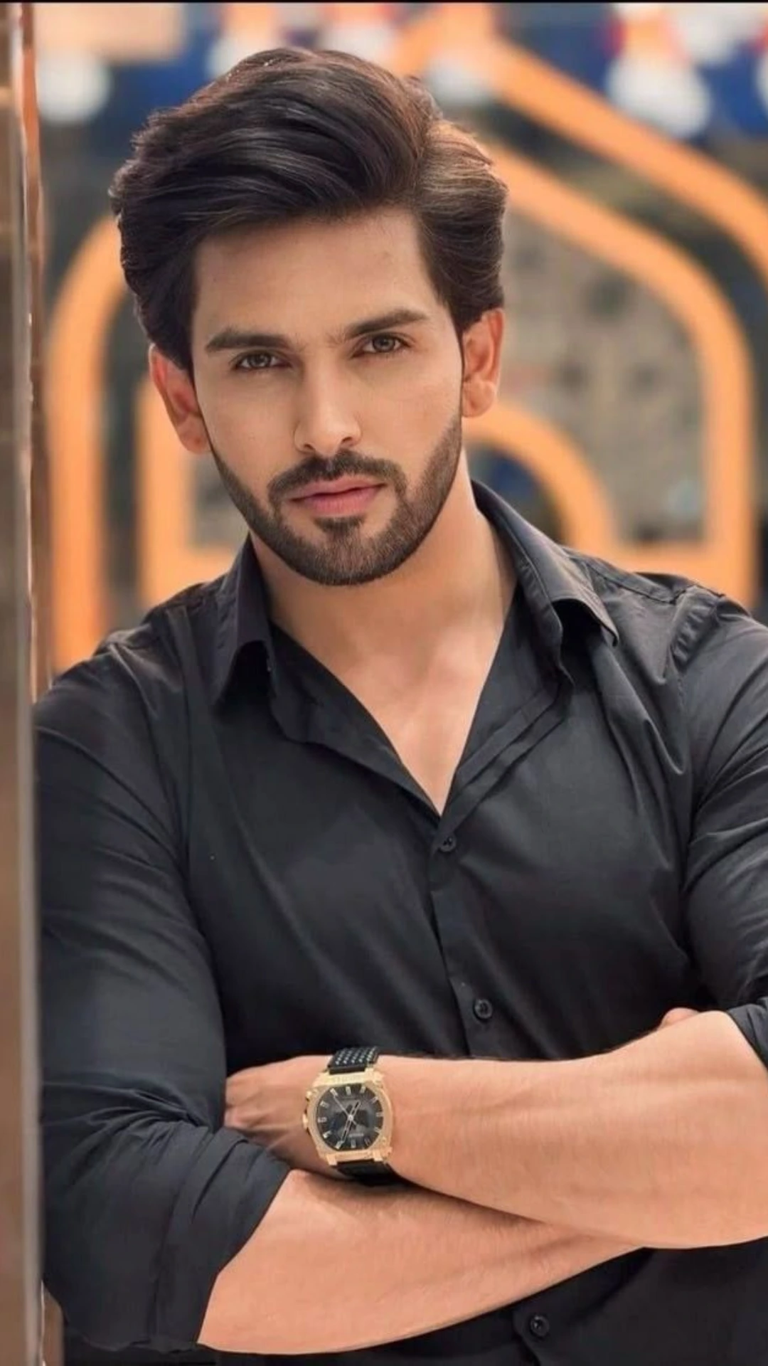 Bearded man with styled dark hair wearing a black shirt and wristwatch, standing with crossed arms against an abstract orange-and-gray background.