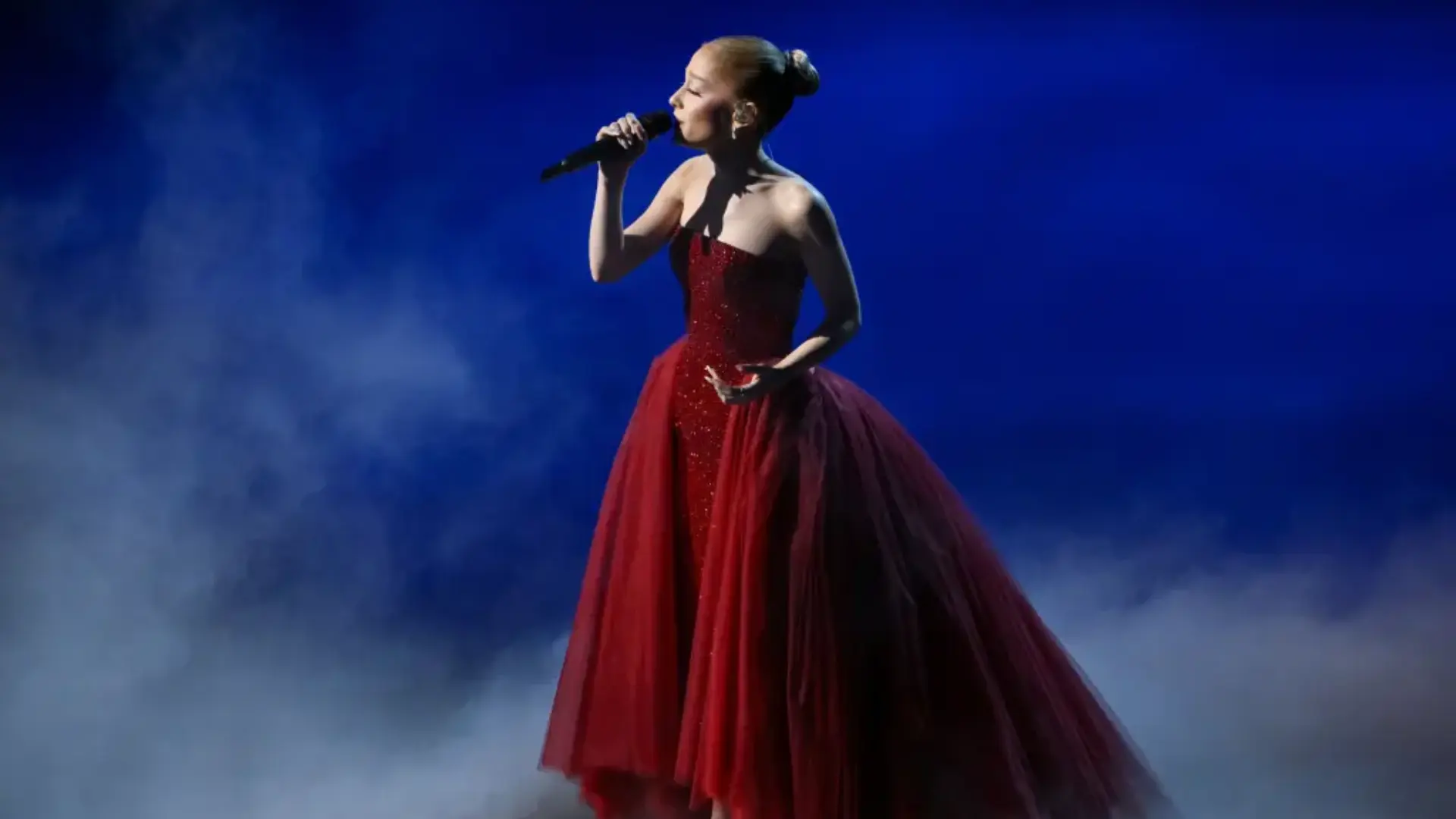 Singer in a strapless red ball gown performs into a handheld microphone on a smoky stage lit with blue light.
