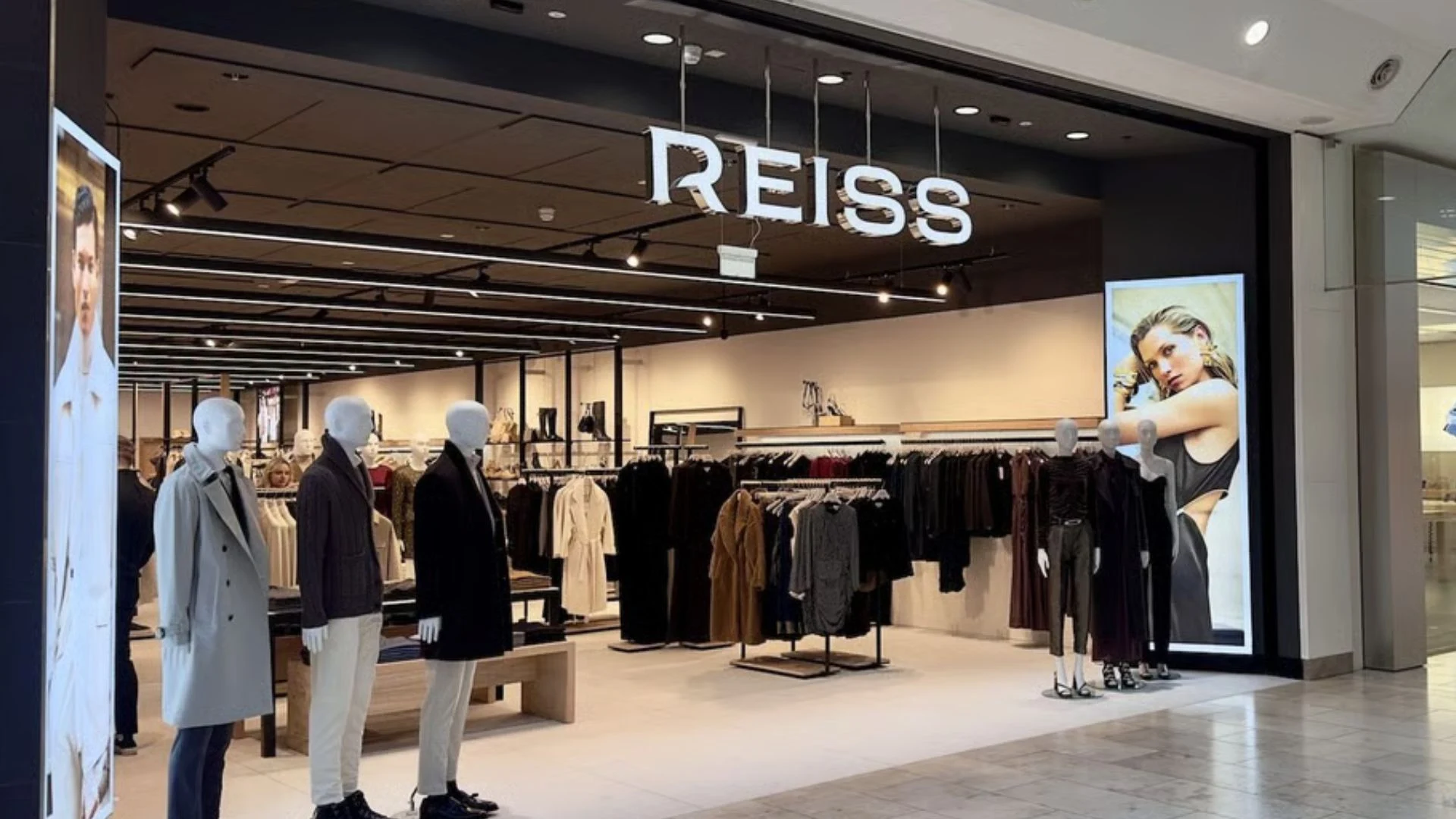 REISS clothing store entrance displaying mannequins in contemporary outfits, clothing racks, and illuminated fashion advertisements in a shopping mall.