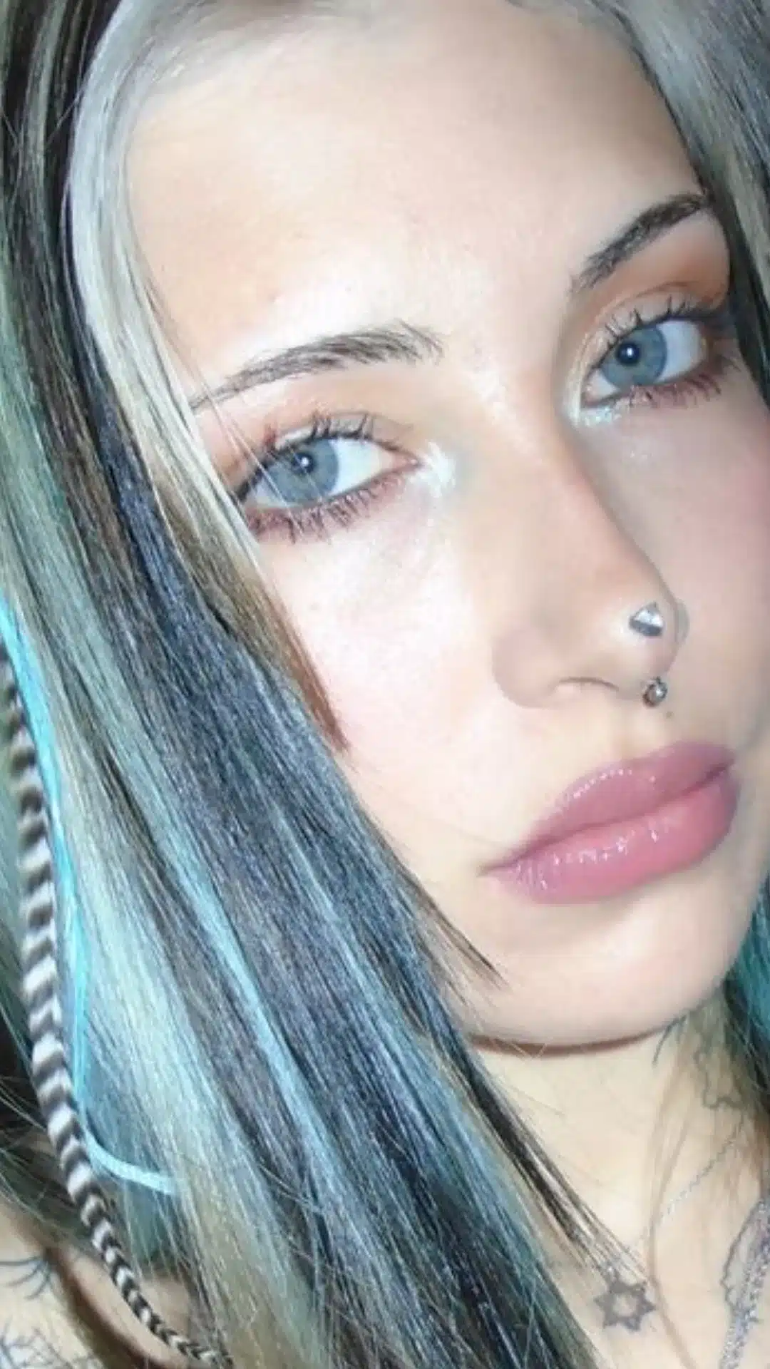 Close-up portrait of a woman with blue eyes, long highlighted hair, glossy pink lips, and nose and lip piercings.