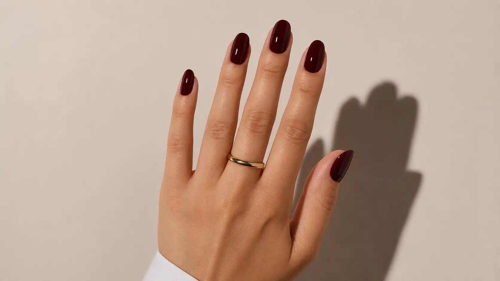Glossy burgundy almond nails displayed on a hand with a simple gold ring against a warm beige background