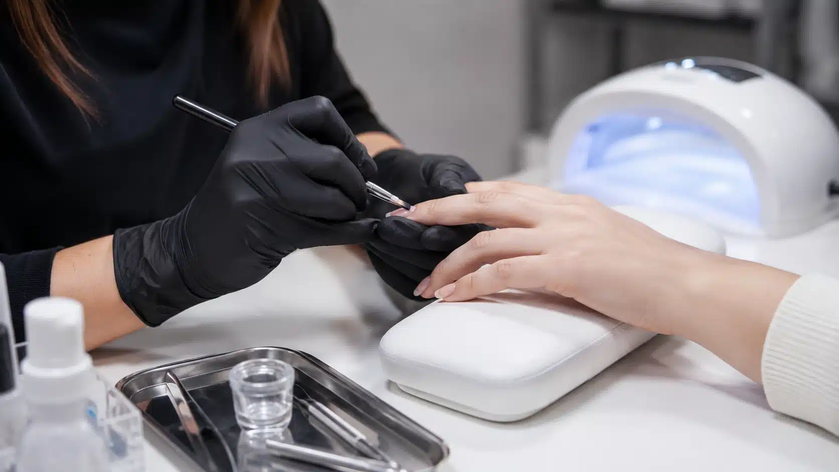 nail technician wearing black gloves carefully applies gel to a client’s fingernail beside a UV lamp and clean tools