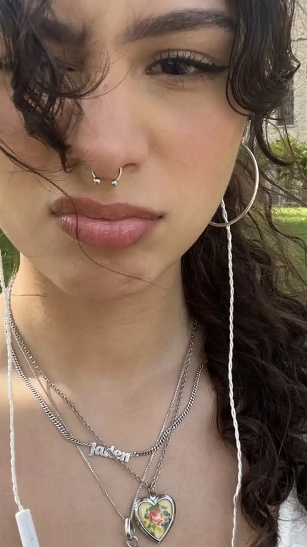 Close-up portrait of a person with curly dark hair, winged eyeliner, a septum ring, hoop earrings, and layered necklaces including a nameplate and heart-shaped pendant.