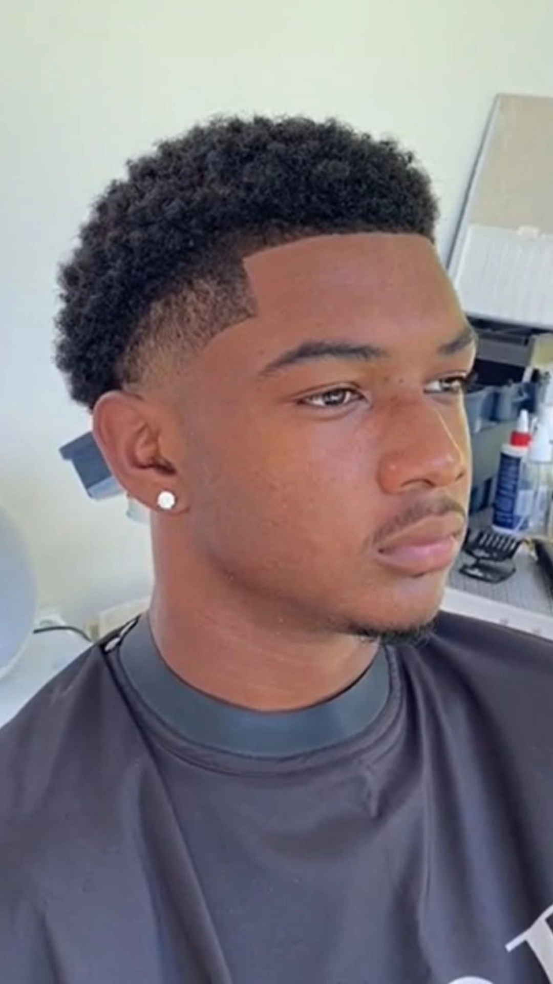 Young man with a fresh low fade haircut and defined hairline wearing a black barber cape in a barbershop.