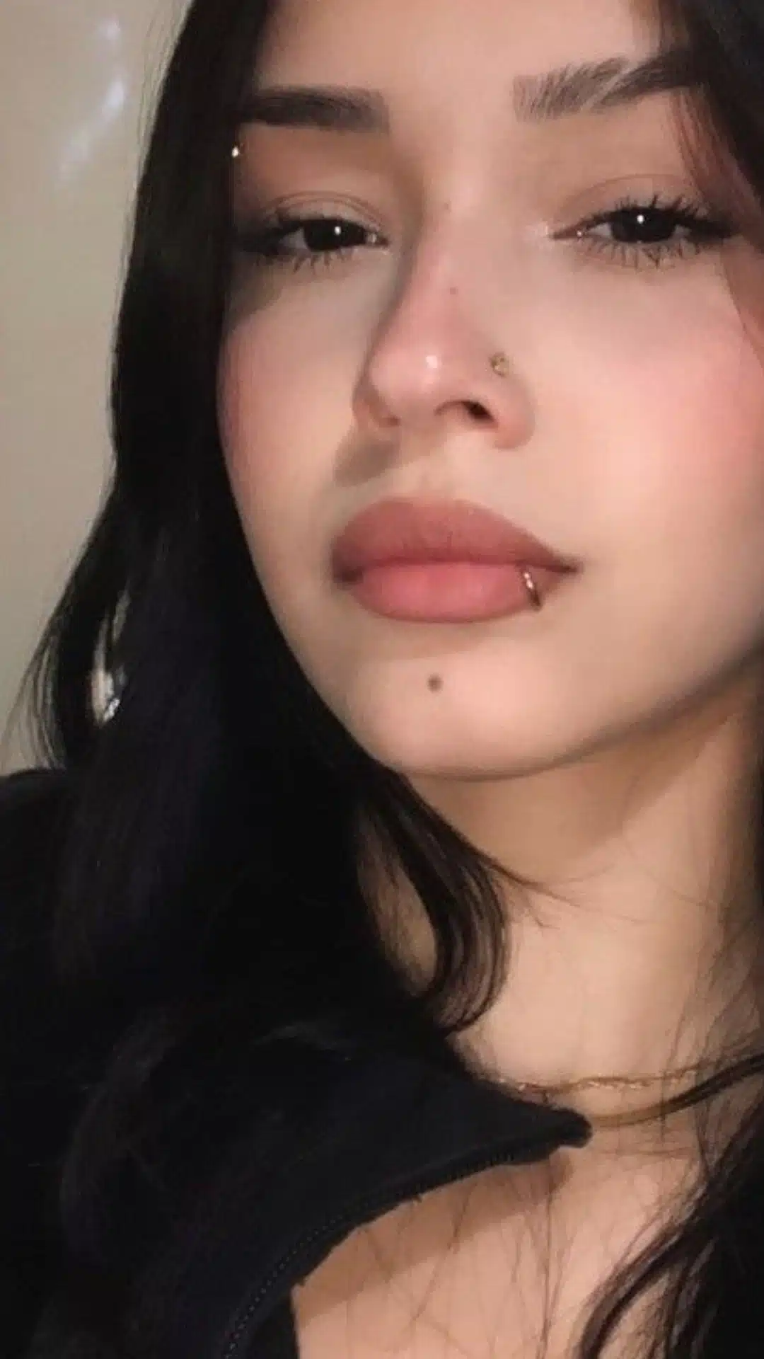 Close-up portrait of a person with dark hair wearing an eyebrow piercing, a nostril stud, and a side labret ring on the lower lip.