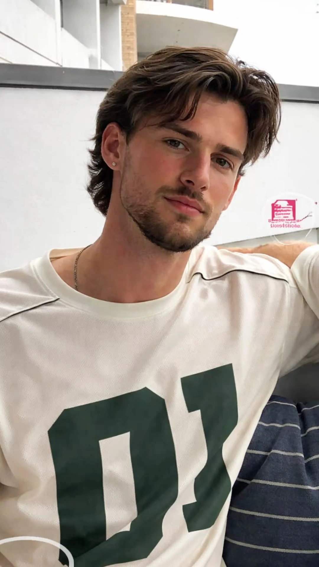 Young man with tousled brown hair and light stubble wearing a white graphic T-shirt, seated indoors against a white wall.