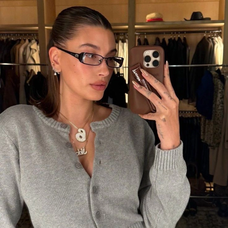 Hailey Bieber wearing slim rectangle glasses taking a selfie in front of a closet, showcasing her stylish eyewear.