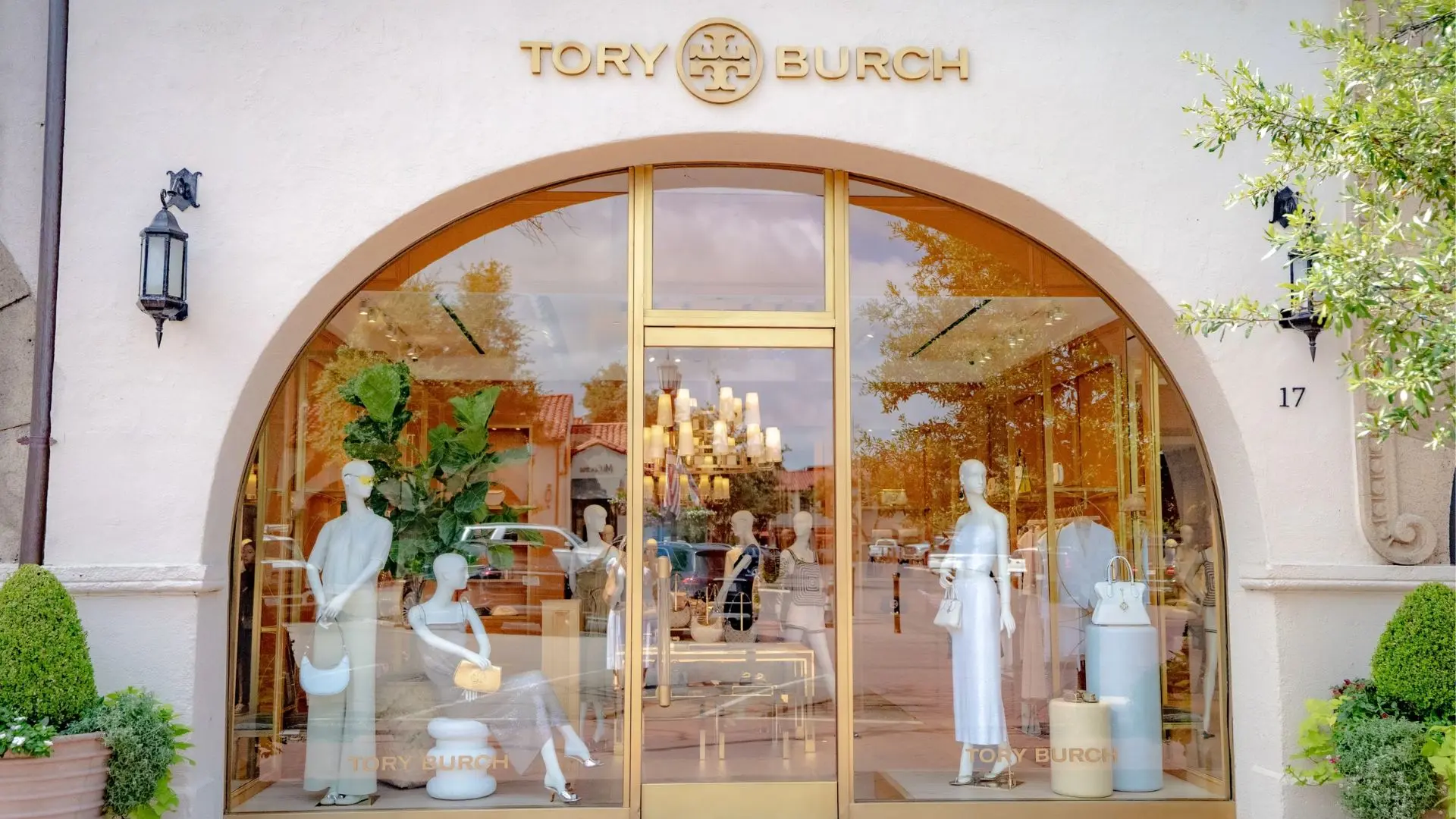 Tory Burch boutique storefront with arched glass doors, mannequins, handbags, gold signage, and landscaped topiary plants.