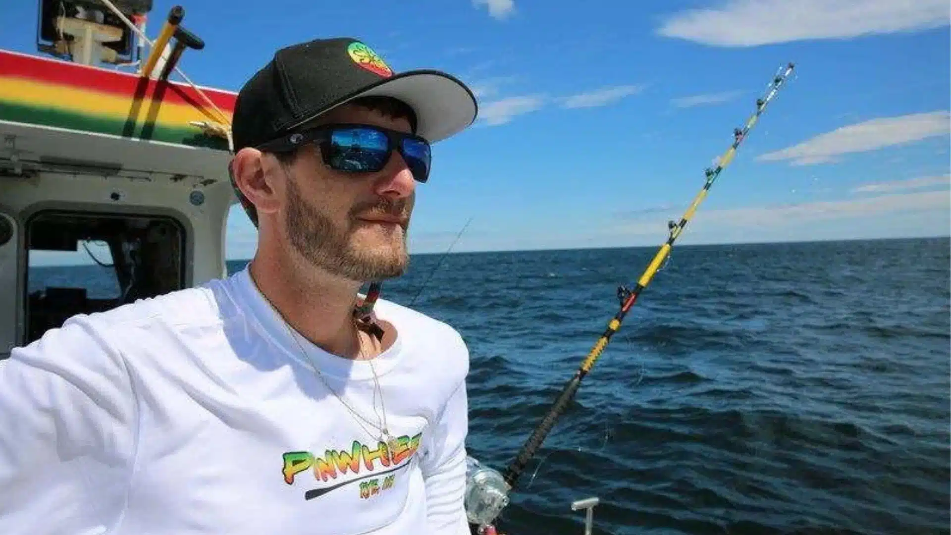 Tyler McLaughlin standing on the PinWheel fishing boat with the ocean and fishing rod in the background under a clear blue sky