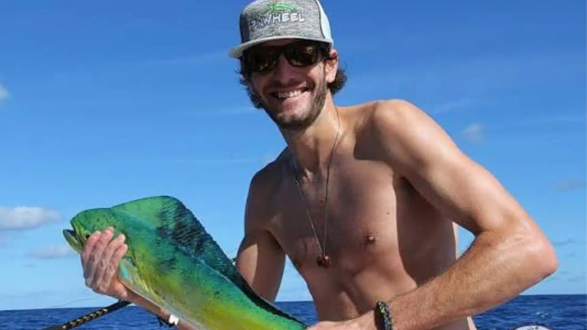 Tyler McLaughlin holds a vibrant dolphinfish aboard a fishing boat on the open ocean under a clear blue sky