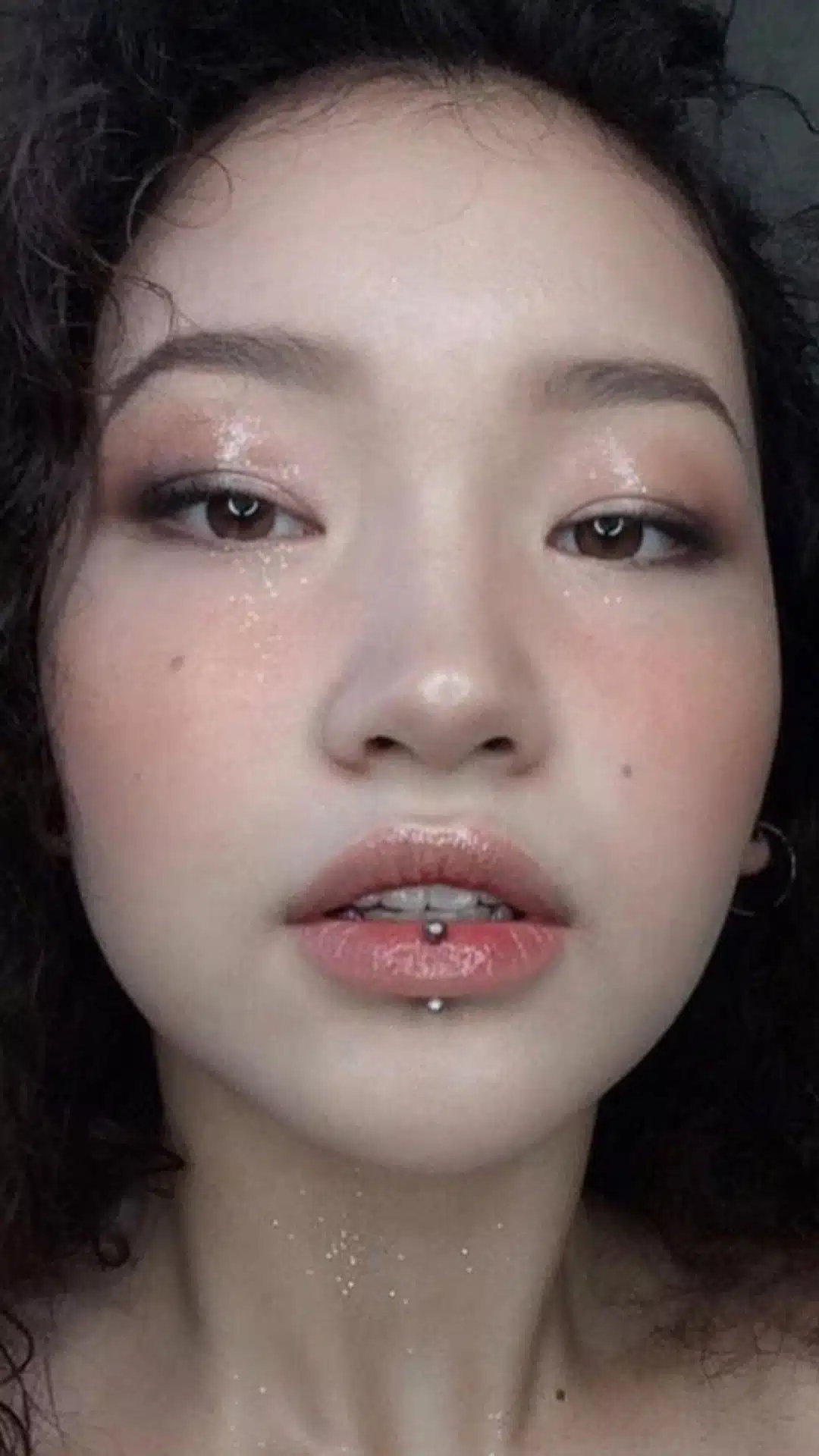 Close-up portrait of a person with dewy pink makeup, glossy lips, dark curly hair, and a lower-lip stud piercing.