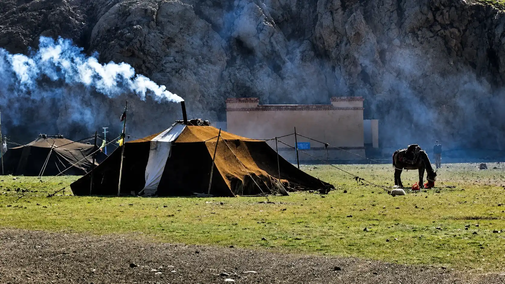 traditional nomad tents set on an open grassland with smoke rising rocky cliffs and a grazing horse nearby under daylight