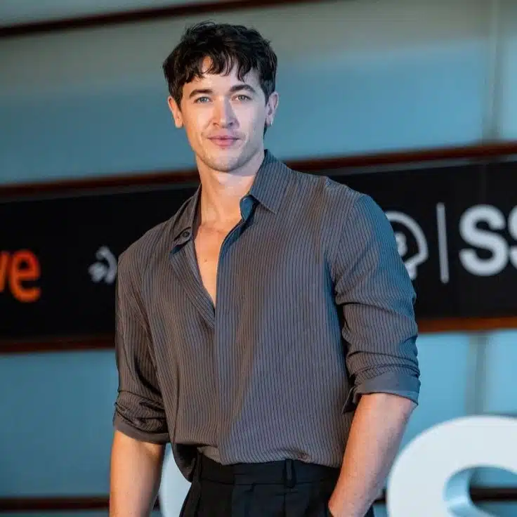 Tom Blyth wearing a grey shirt and black pants stands confidently, showcasing a casual yet polished appearance.