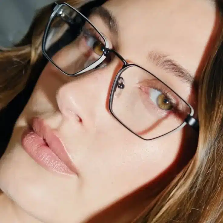 Hailey Bieber wearing wire rim glasses and a black shirt, posing confidently for the camera.