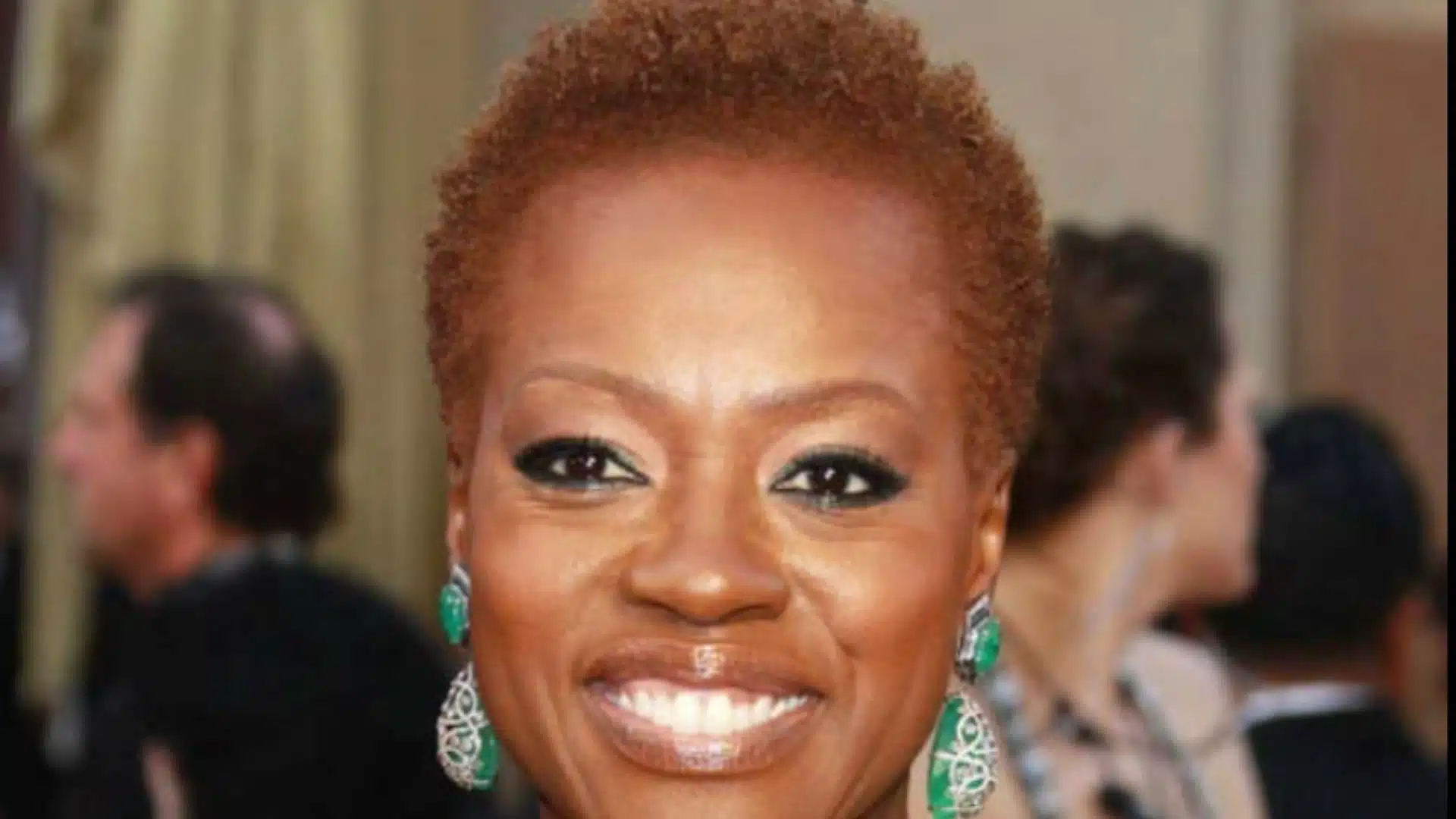 woman with short auburn hair smiles at a formal event wearing green statement earrings and dramatic dark eye makeup