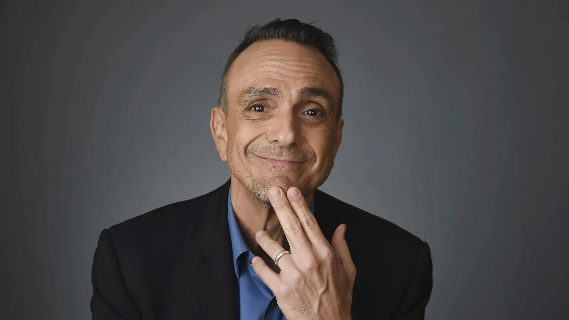 Hank Azaria smiling with a hand touching his chin against a gray backdrop.