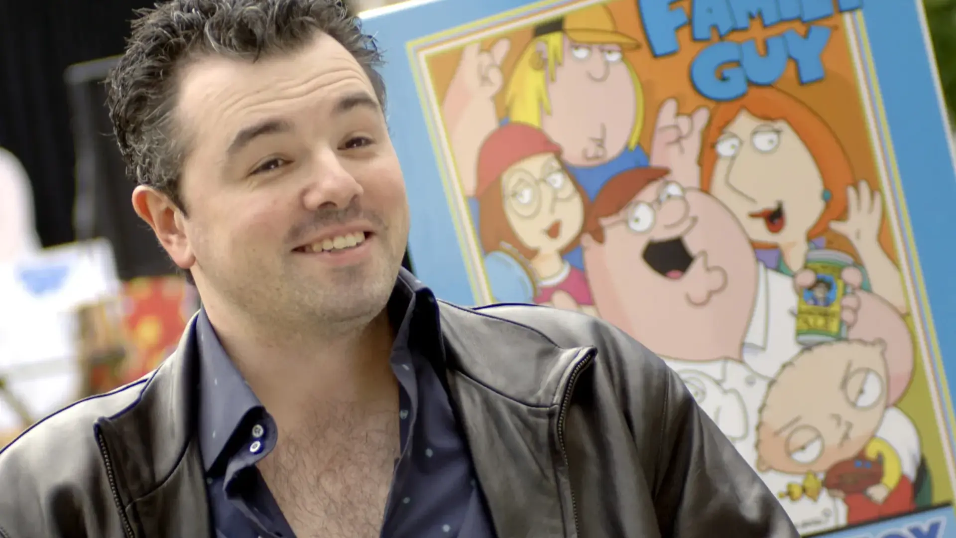 Seth MacFarlane smiling next to a Family Guy poster